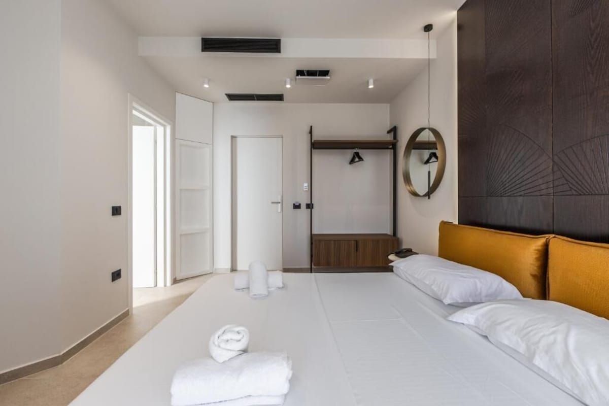 Ein Hotelzimmer mit einem Bett, einem Spiegel, einer hölzernen Bank und einer Tür zu einem weiteren Raum.