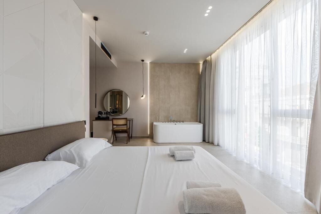 A hotel room with a bed, a desk, a mirror, a bathtub, and large windows.