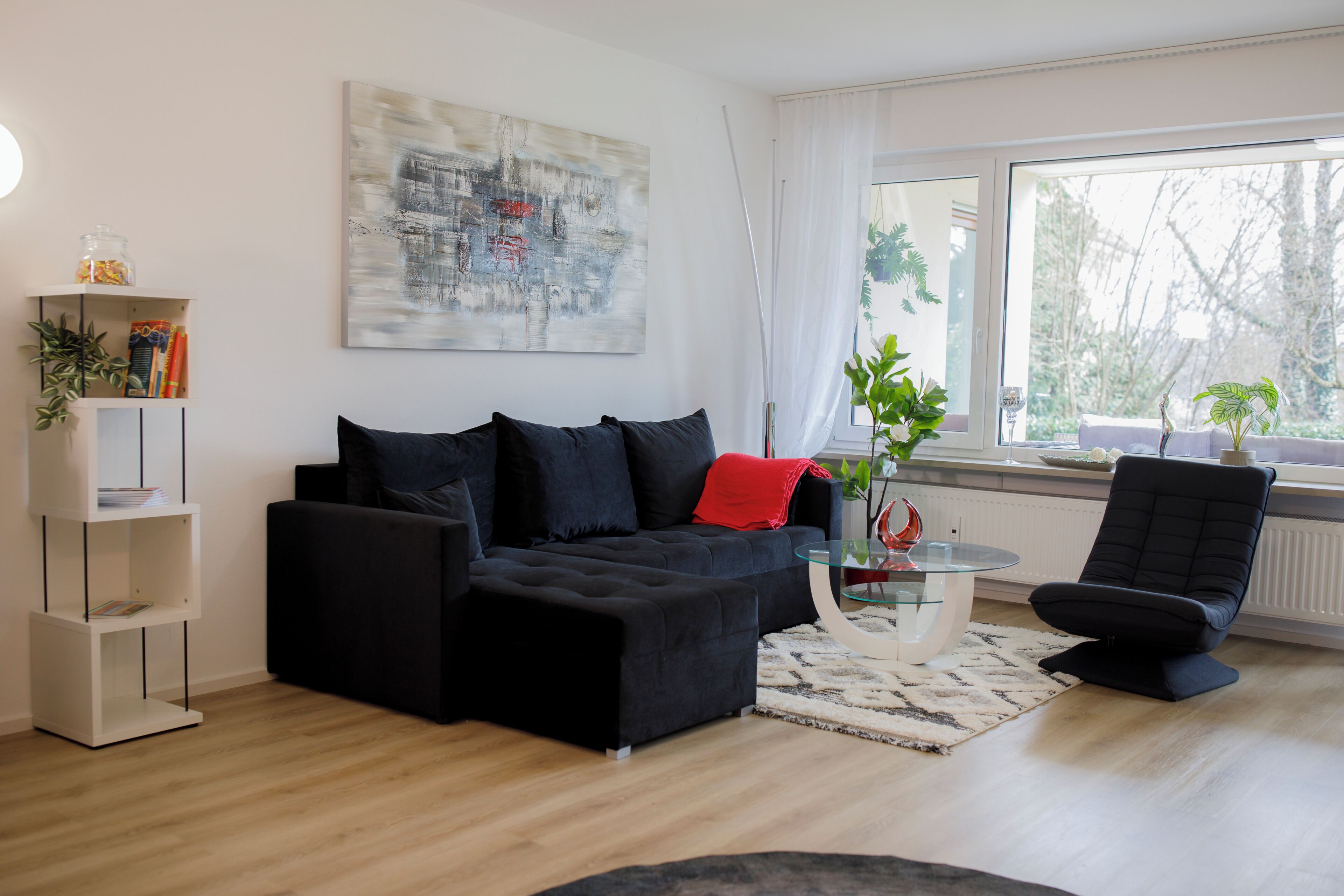 Ein modernes Wohnzimmer mit einer schwarzen Couch, einem Glastisch und einem großen Fenster mit Blick auf Bäume.