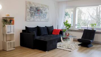 Ein modernes Wohnzimmer mit einer schwarzen Couch, einem Glastisch und einem großen Fenster mit Blick auf Bäume.