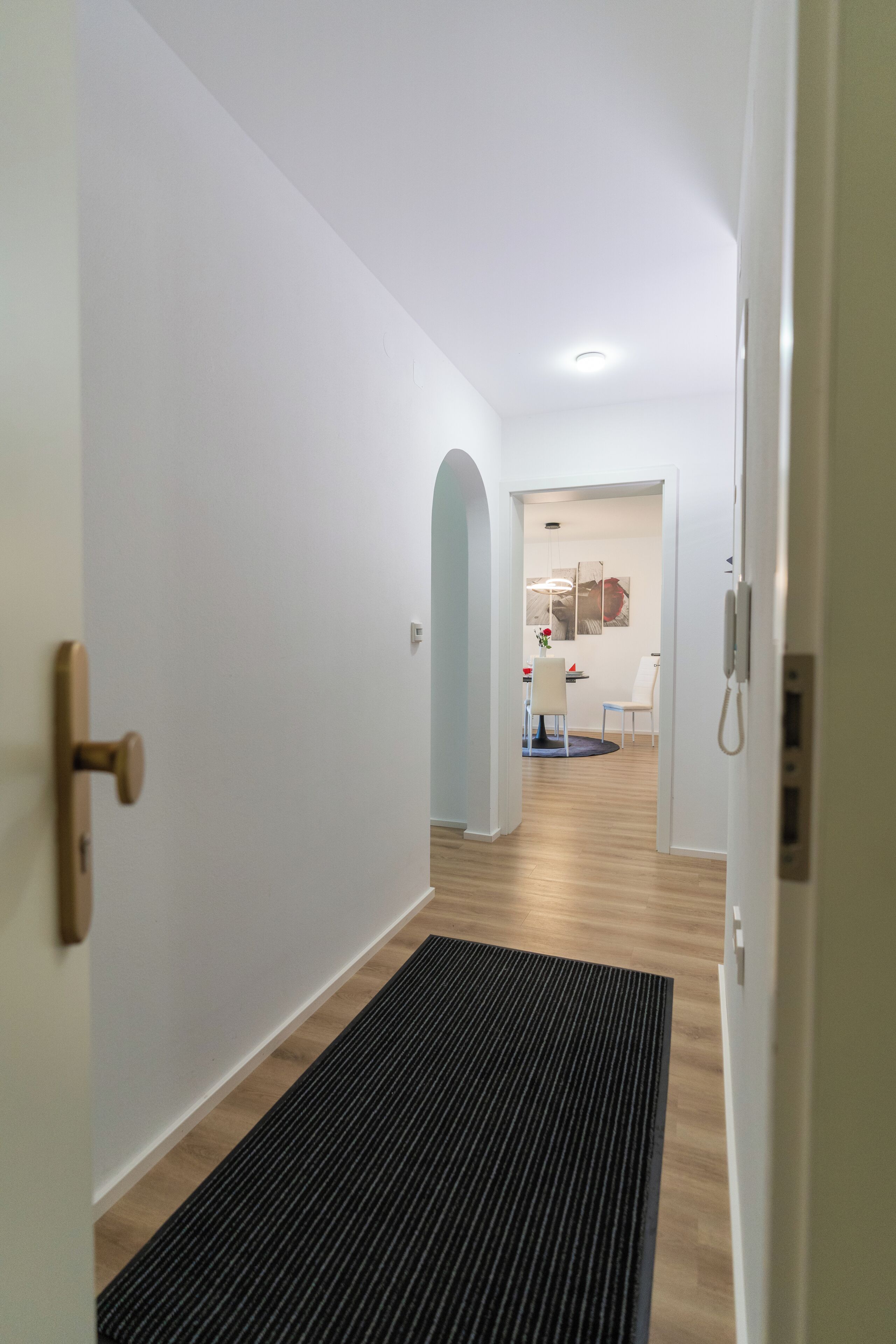 A hallway with a black mat, white walls, and a wooden floor.