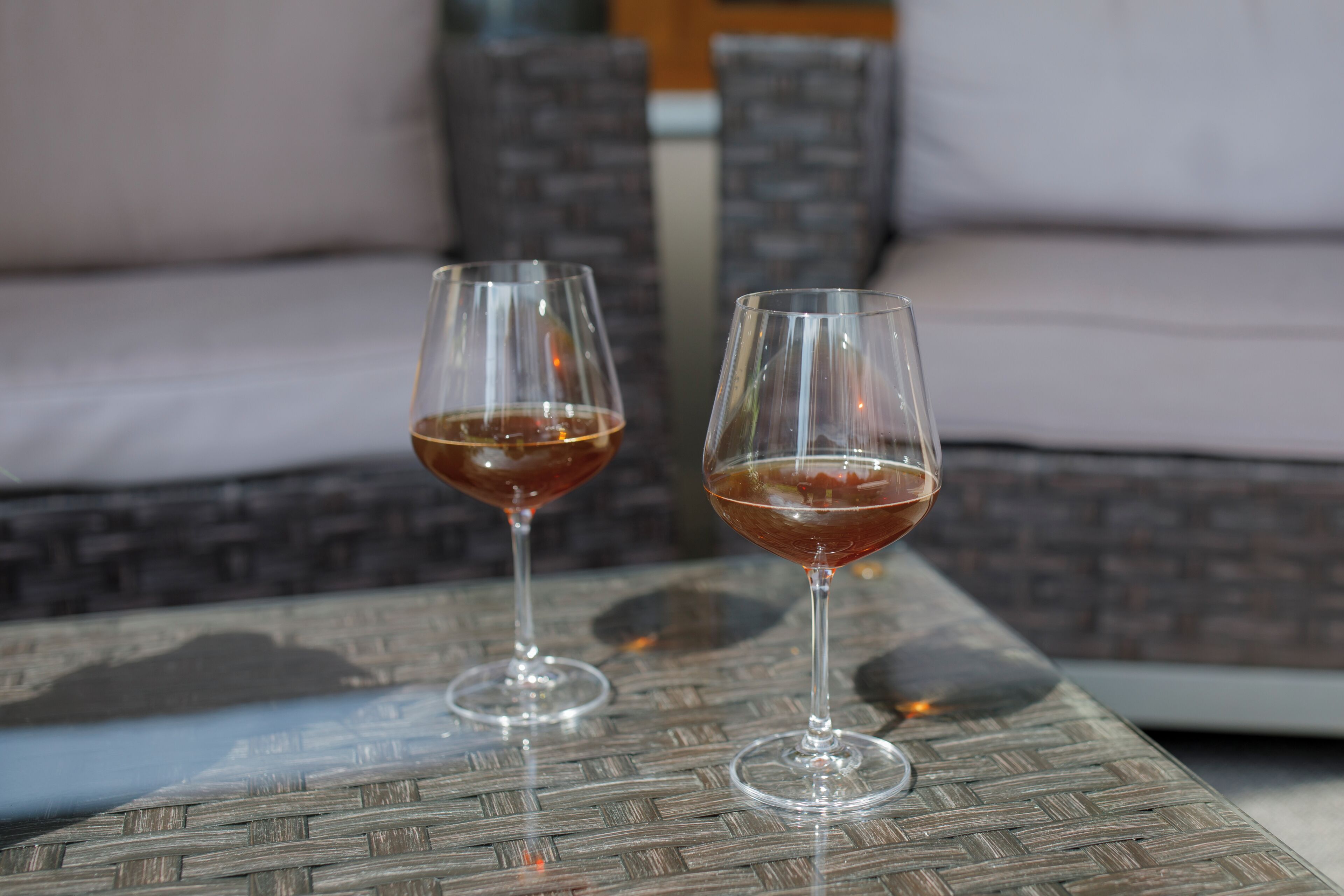 Two wine glasses on a woven table with a sofa in the background.