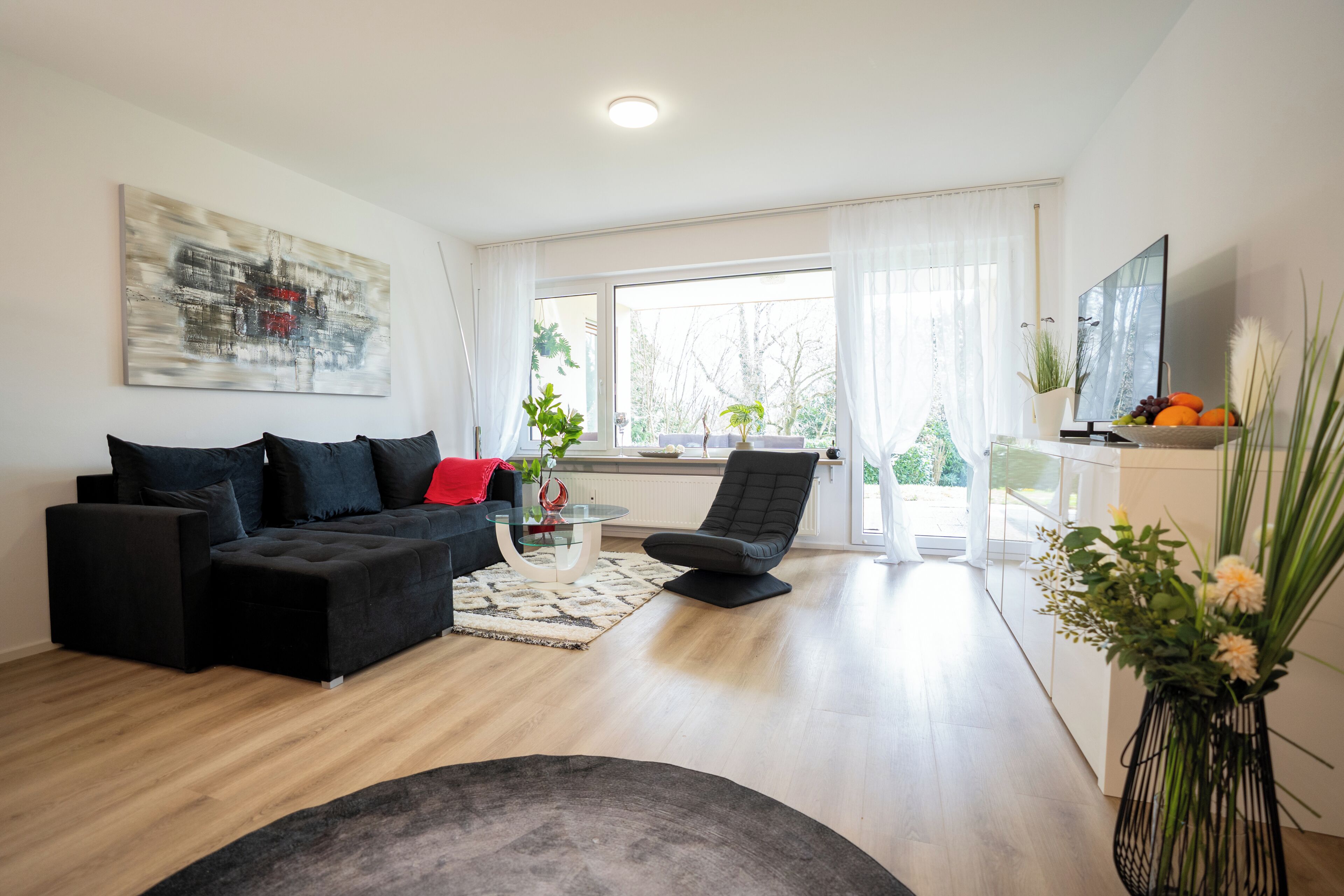 Ein modernes Wohnzimmer mit einem schwarzen Sofa, einem Glastisch und einem schwarzen Sessel. Ein großes Fenster mit durchsichtigen Vorhängen, ein Gemälde an der Wand und eine Vase mit Blumen auf einem Beistelltisch.