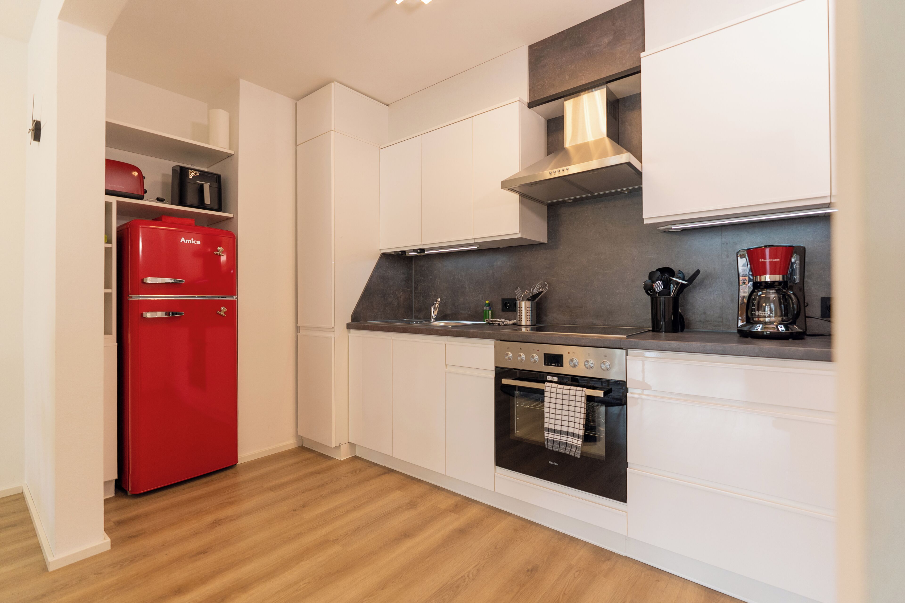 Eine moderne Küche mit weißen Schränken, einem roten Amica-Kühlschrank, einer Edelstahl-Dunstabzugshaube, einer Kaffeemaschine und einem Backofen.