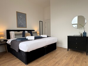A bedroom with a black bed, white bedding, a bedside table with a lamp, a dresser, a mirror, and a framed artwork on the wall. - 2-Bed, 2-Bath Near Birkdale Golf Club - Flat 5 (Southport)