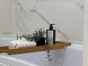 A bathroom with a marble wall, a wooden shelf with a towel and a bottle of Fiori di Bach body lotion, and a sprig of rosemary. - 2-Bed, 2-Bath Near Birkdale Golf Club - Flat 5 (Southport)