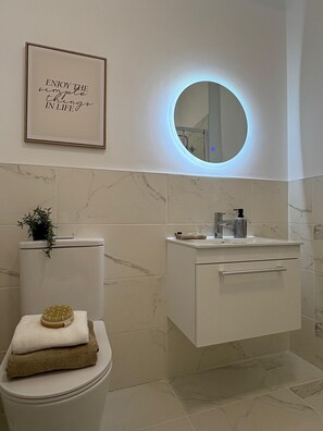 A modern bathroom with a toilet, a sink, and a mirror with a circular light. There is a framed quote on the wall and a plant on the toilet. - 2-Bed, 2-Bath Near Birkdale Golf Club - Flat 5 (Southport)