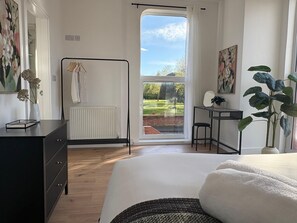 A bedroom with a bed, a radiator, a desk, a chair, and a large window with a view of a garden. - 2-Bed, 2-Bath Near Birkdale Golf Club - Flat 5 (Southport)