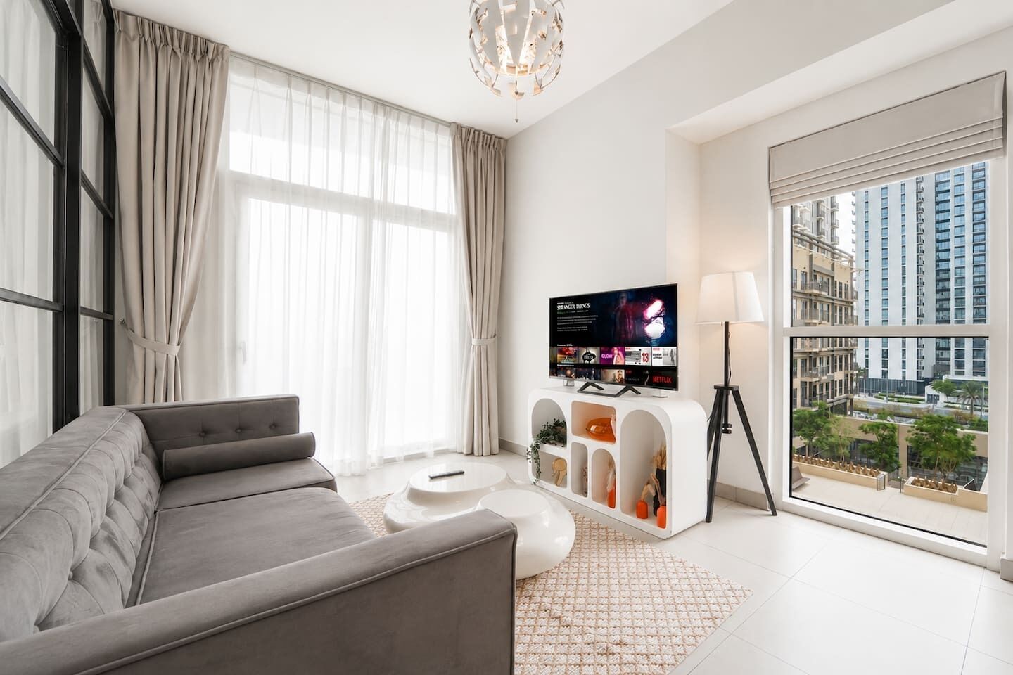 Un salon moderne avec un canapé gris, un téléviseur à écran plat, une table basse blanche et une vue sur le paysage urbain, visible à travers de grandes fenêtres.