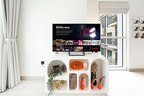 A modern living room with a flat-screen TV displaying 'Stranger Things' on Netflix, a white entertainment unit with decorative items, and a view of a building through a large window. - Bright & Stylish 2BR Retreat | Socio Dubai Hills (Dubai)