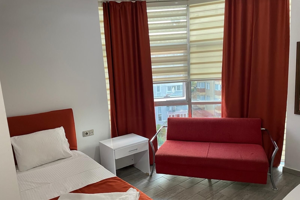 A hotel room with a bed, a red sofa, a small white table, and a window with curtains.