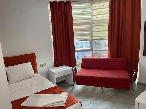 A hotel room with a bed, a red sofa, a small white table, and a window with curtains.