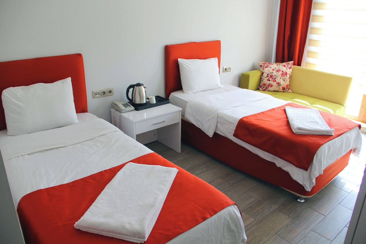 A hotel room with two single beds, a nightstand with a kettle, a telephone, and a small table.