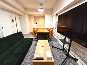 A compact living space with a green sofa, a wooden table with remote controls, and a view of a kitchen area. - TOWER HOTEL (Fukuoka)