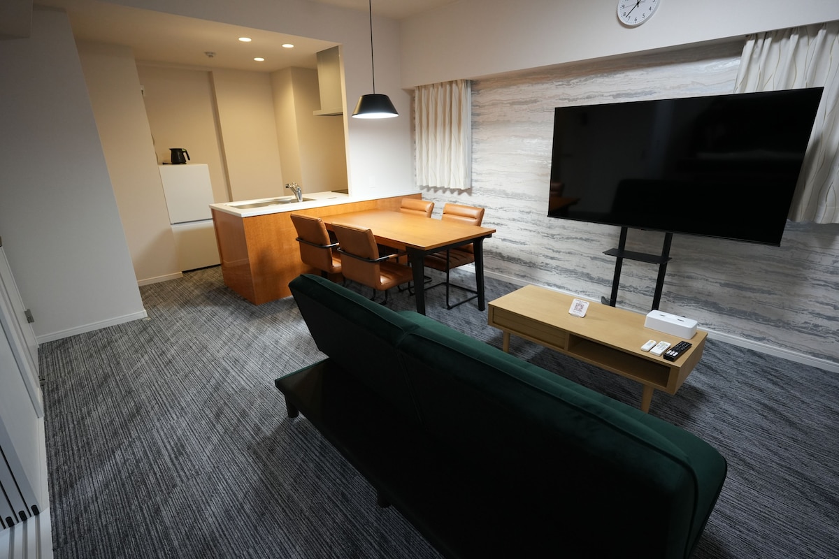 A hotel room with a sofa, a dining table, a TV, and a kitchen area.