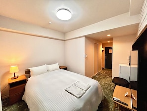 Quarto de hotel com cama, mesa de cabeceira, luminária e escrivaninha com cadeira.