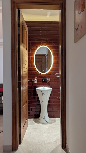 Une salle de bain avec un lavabo en marbre blanc, un miroir rond avec un éclairage intégré et une porte en bois.