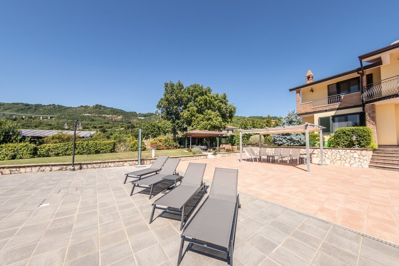 A rooftop terrace with sun loungers, a pergola, and a view of a hill.