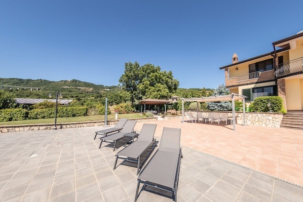 A rooftop terrace with sun loungers, a pergola, and a view of a hill.