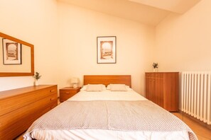 A bedroom with a bed, a nightstand, a dresser, a radiator, and two framed pictures on the wall.
