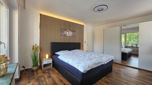 A modern bedroom with a wooden slat wall, a bed with floral bedding, a bedside table with a lamp, and a view of the outdoors through a window. - Exclusive, bright apartment with balcony in the city, 1 Gbit Wifi, boxspringbeds (Kassel)