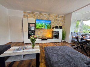 A modern living room with a flat-screen TV mounted on a stone wall, a dining area with a table and chairs, and a sofa. - Exclusive, bright apartment with balcony in the city, 1 Gbit Wifi, boxspringbeds (Kassel)