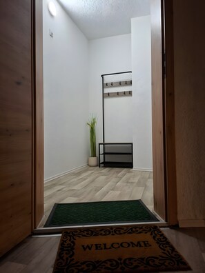 A room with a wooden floor, a green doormat with 'WELCOME' written on it, a small potted plant, and a black metal shelf with hooks.