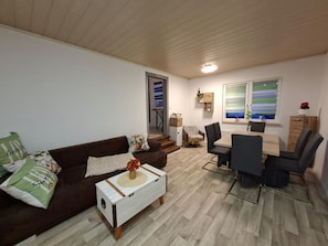 Een woonkamer met een bruine bank, een witte salontafel en een eethoek met een tafel en stoelen.