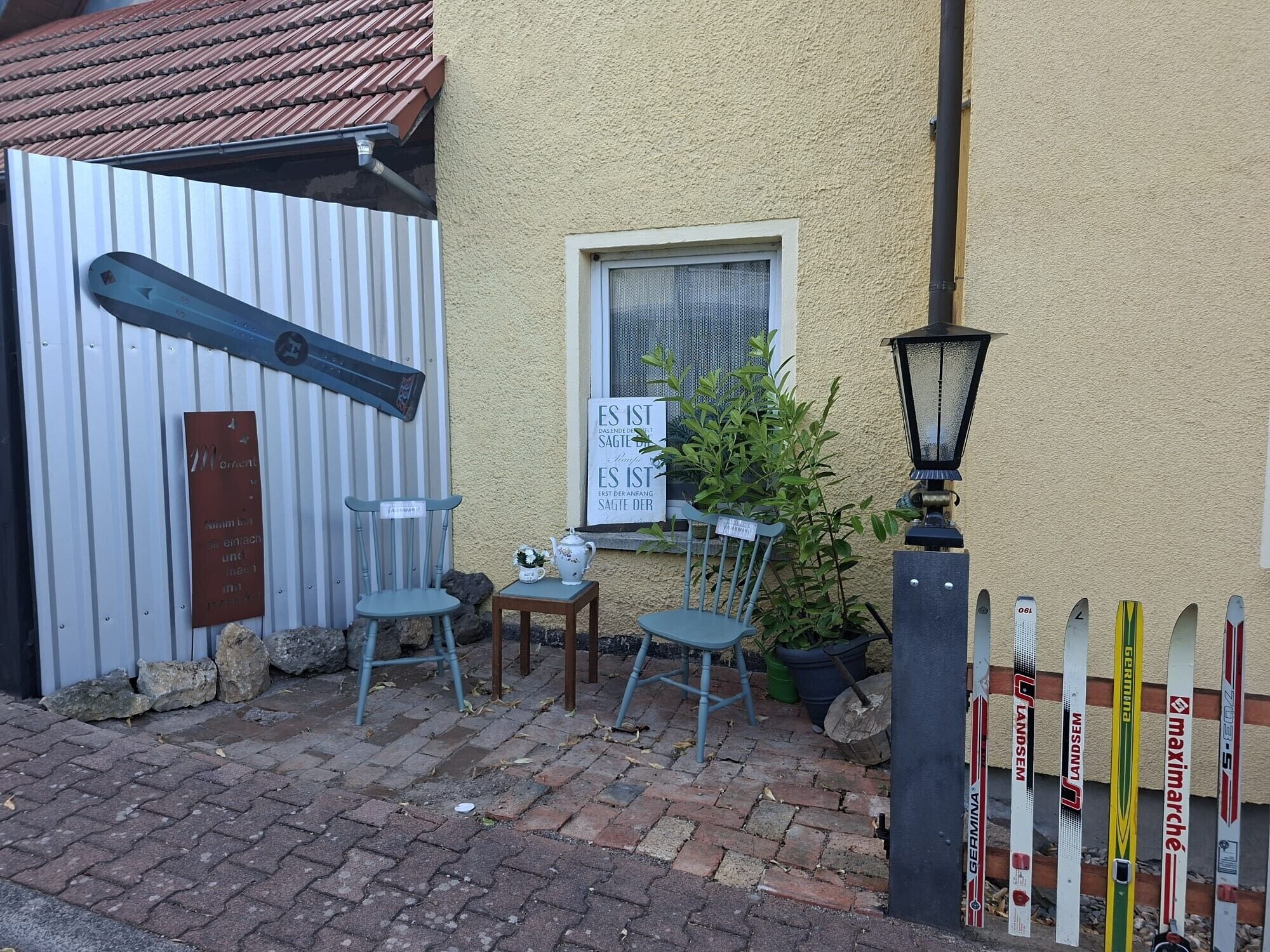 A small outdoor seating area with two chairs and a table, a sign with German text, and a row of skis leaning against a wall.