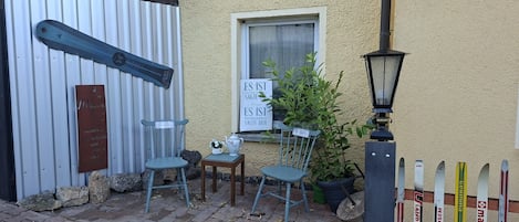 Un petit espace extérieur aménagé avec deux chaises et une table, une enseigne avec du texte en allemand, et une rangée de paires de skis appuyés contre un mur.