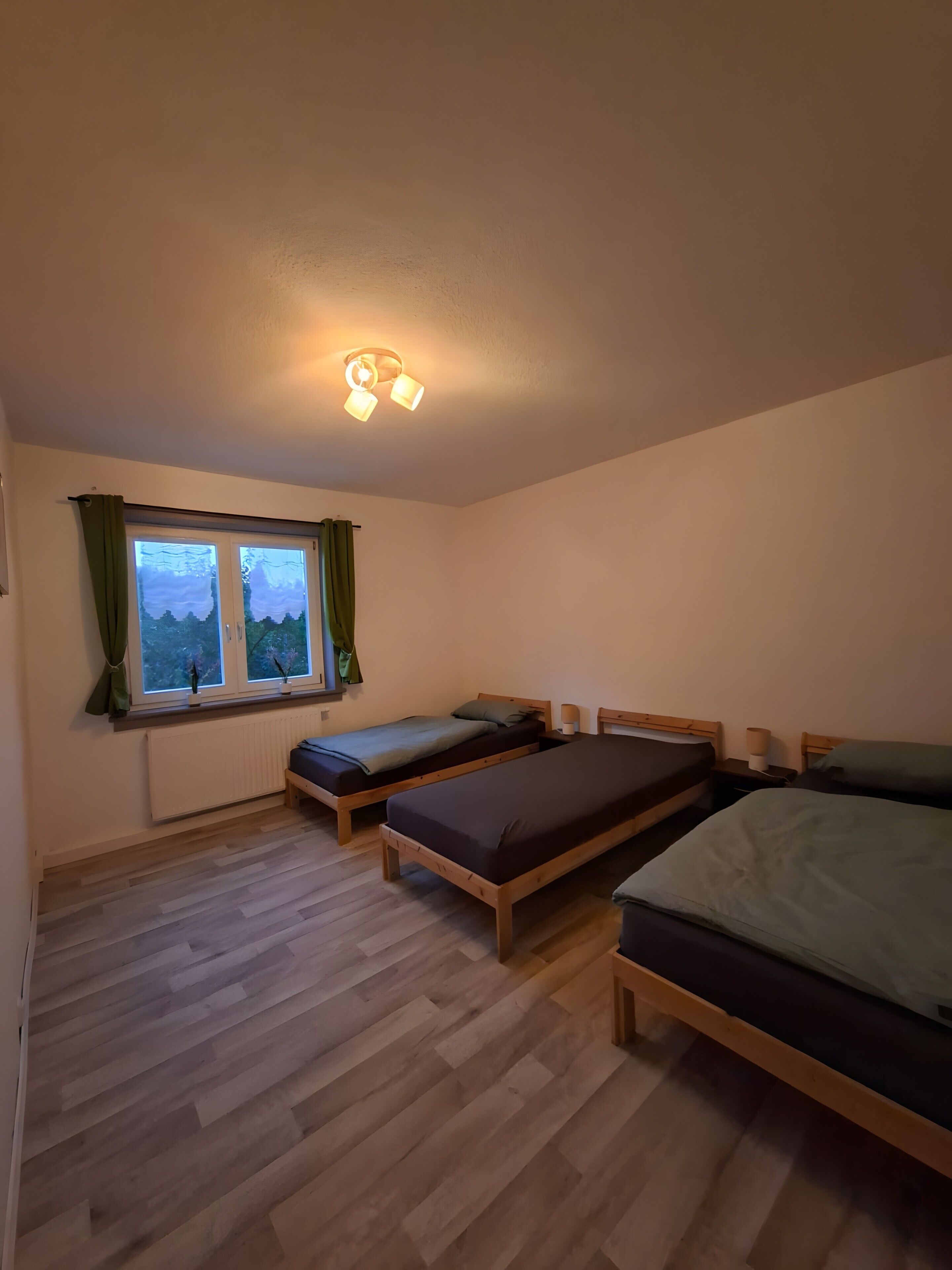 A room with two wooden beds, a window with curtains, a radiator, and a ceiling light fixture.
