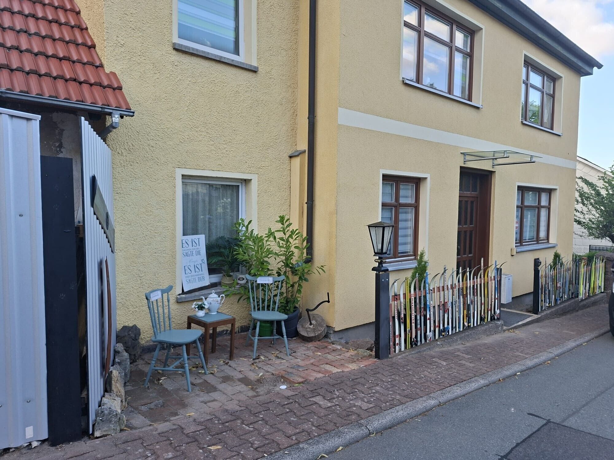 A residential building with a yellow exterior, a paved driveway, a small outdoor seating area with a table and chairs, and a row of skis leaning against a fence.