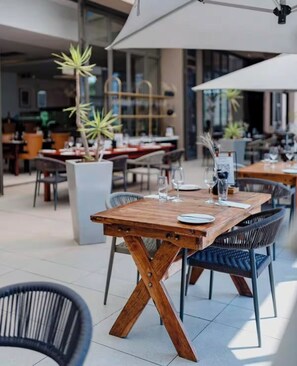 Een terras met houten tafels en stoelen, plantenbakken en een grote parasol voor beschutting.
