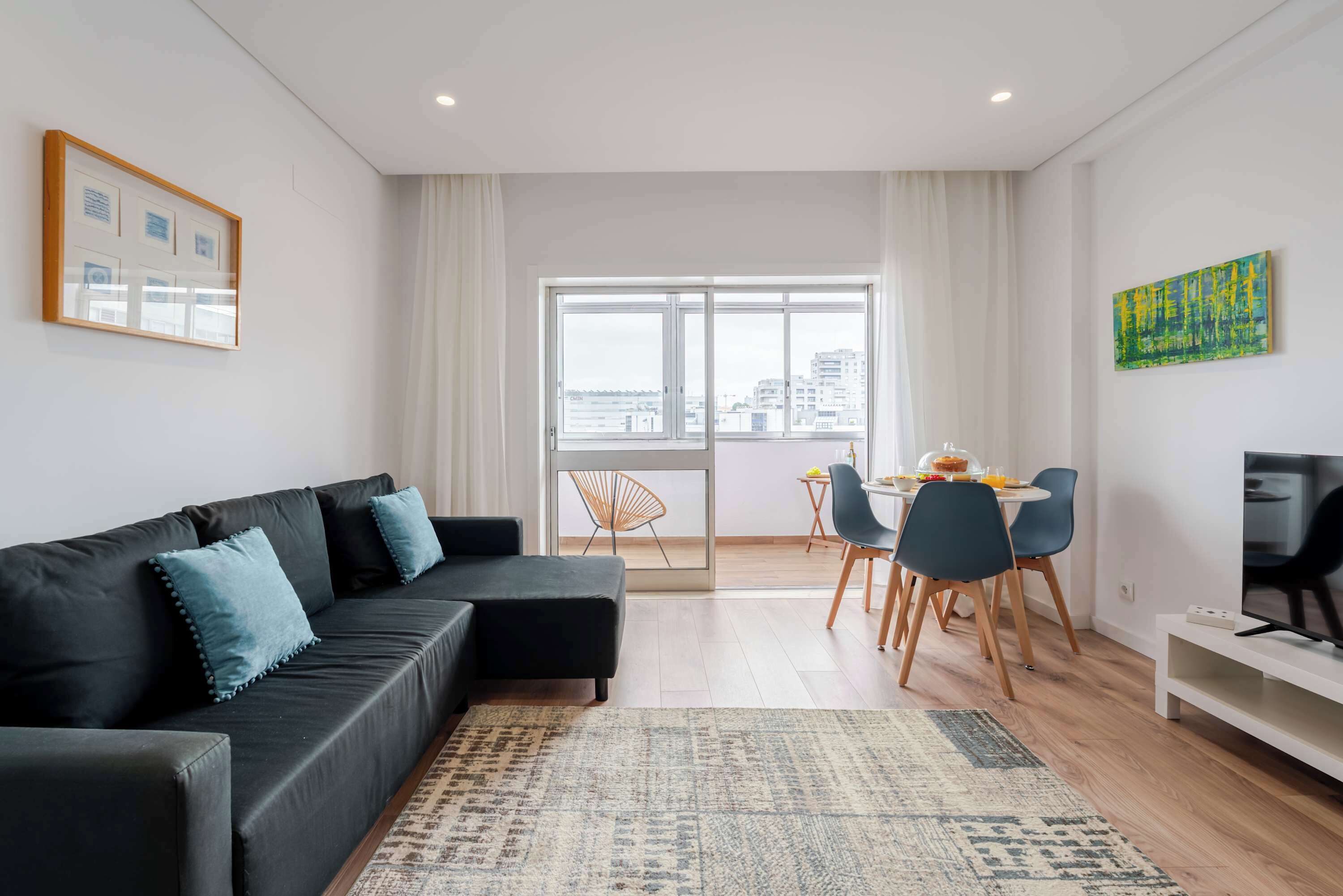 Un salon moderne avec un canapé d’angle noir, un coin repas comprenant une table et des chaises, et une grande fenêtre donnant sur la ville.