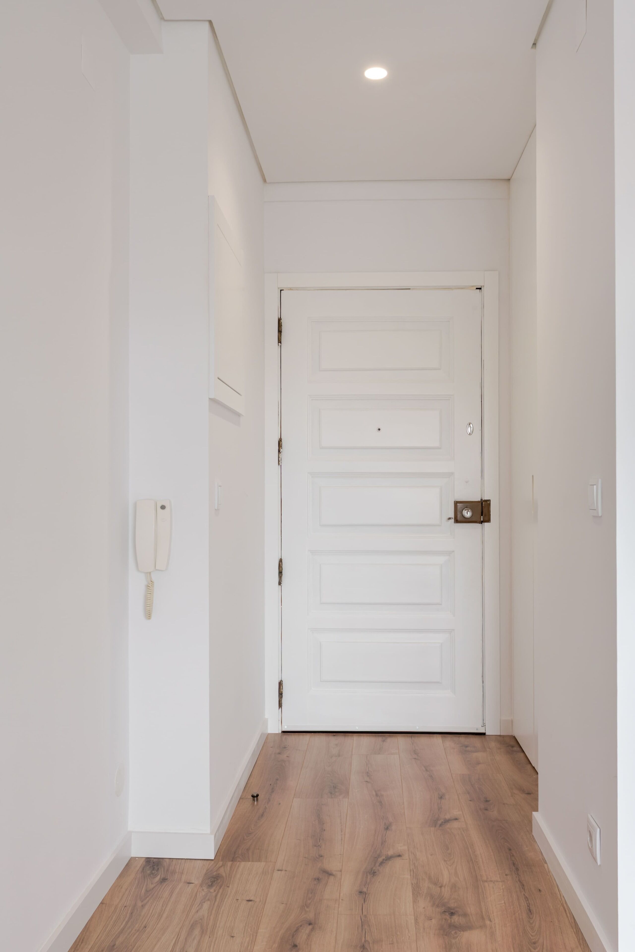Un pasillo con una puerta blanca, suelo de madera y un teléfono fijado a la pared.