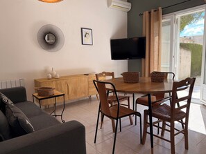 A modern living room with a sofa, a dining table with chairs, a TV mounted on the wall, and a window with curtains. - Le Tournesol - Laforest Estate Vacation Rental (Fontvieille)