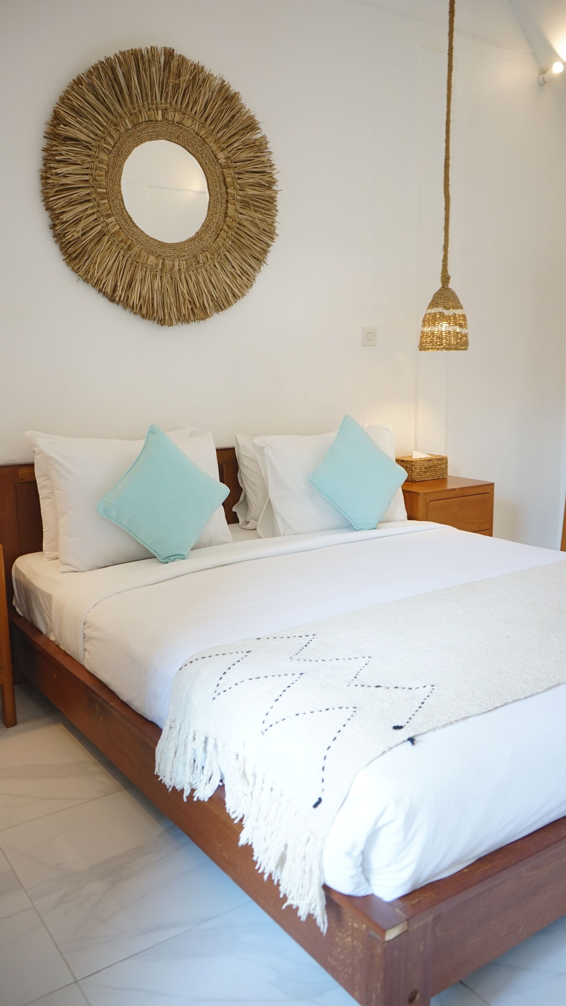 Una cama con ropa de cama blanca y almohadas azules, un adorno de pared tejido, una lámpara de noche y una mesita de noche de madera.