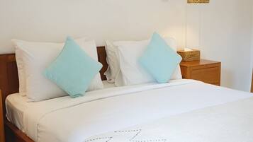 Una cama con ropa de cama blanca y almohadas azules, un adorno de pared tejido, una lámpara de noche y una mesita de noche de madera.
