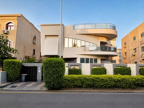 Un edificio moderno de varios pisos con un balcón curvo, entrada protegida por una reja y setos bien cuidados.
