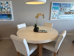 Ein runder Esstisch mit vier weißen Stühlen, eine Topforchidee und ein Buch. An der Wand hängen zwei Bilder, eines zeigt Blumen, das andere eine Strandansicht.