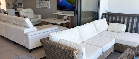 A modern living room with a white sofa set, a wicker accent piece, and a flat-screen TV on a stand.