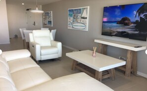 Ein Wohnzimmer mit einem weißen Sofa, einem weißen Sessel, einem Holztisch und einem an der Wand befestigten Flachbildfernseher.