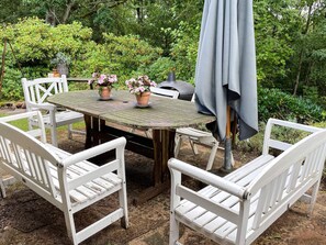 Een houten tafel met een grijze parasol, witte stoelen en potplanten.