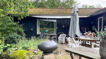 A rustic outdoor seating area with a wooden table and chairs, a large black urn, and a covered patio with a tiled roof.