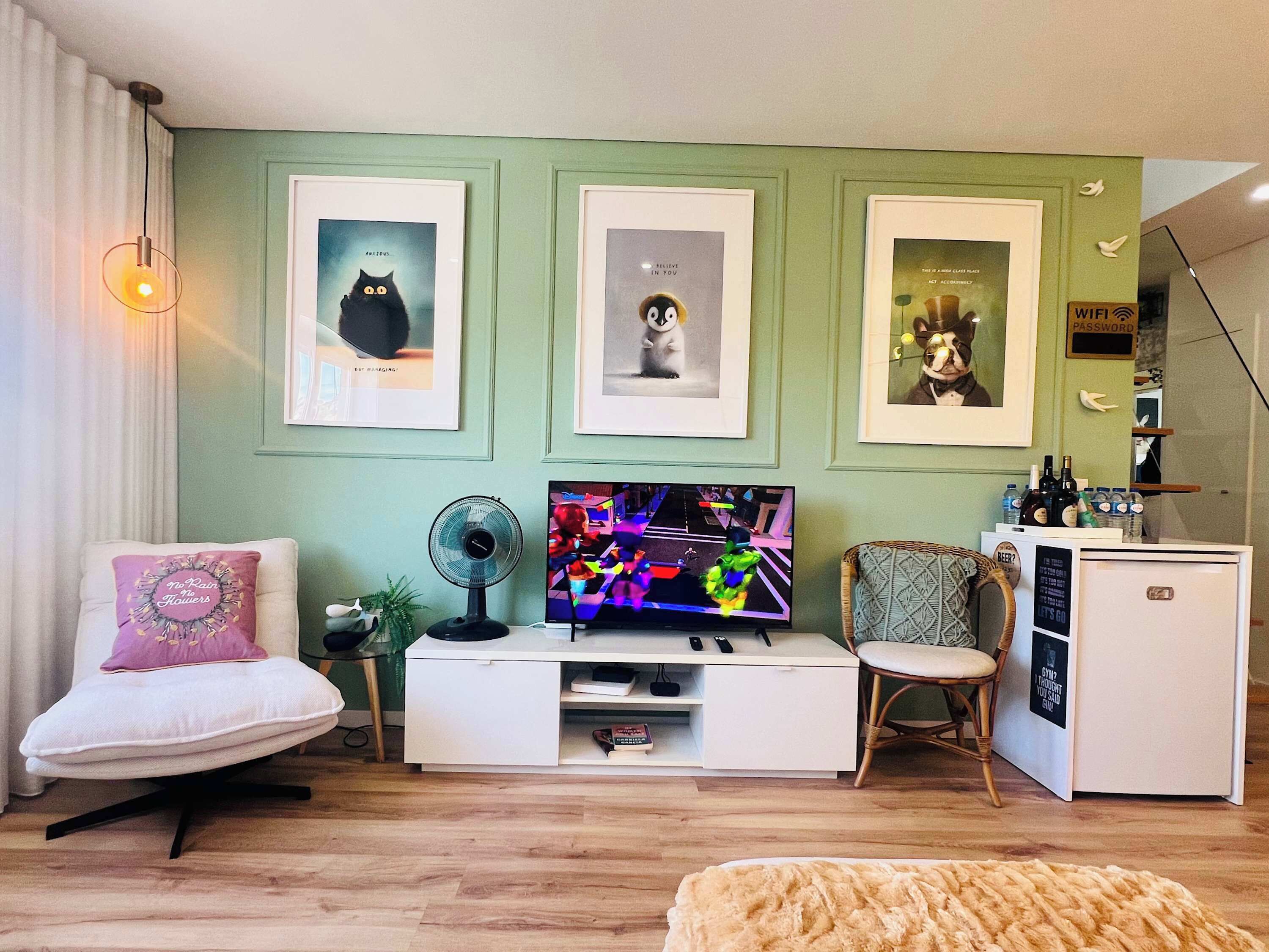 A living room with a TV, framed pictures, a fan, a chair, and a small refrigerator.