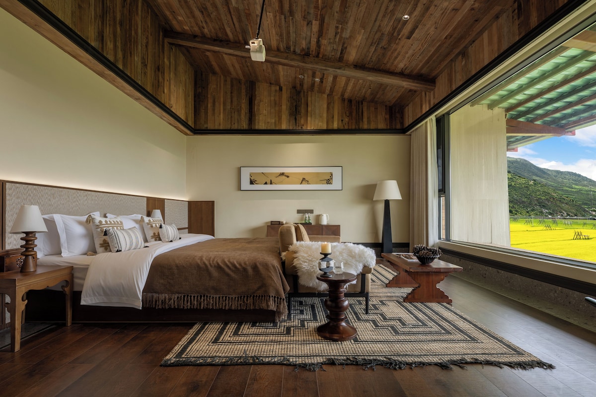Un dormitorio con una cama grande, mesitas de noche, una lámpara, un cuadro en la pared y vistas a un paisaje verde a través de una ventana.