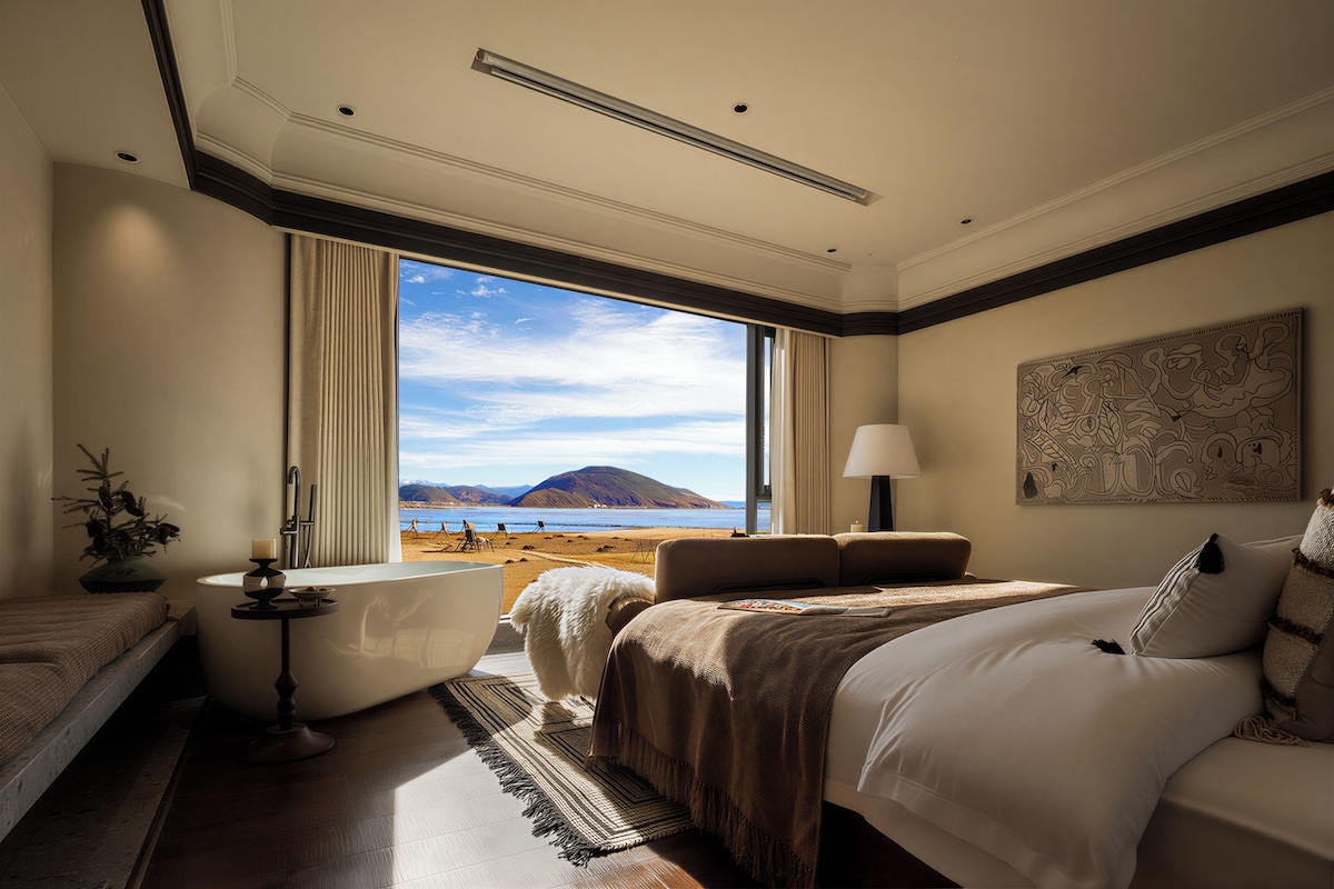 Habitación de hotel con una cama grande, zona de estar, bañera y vistas a la playa y las montañas.
