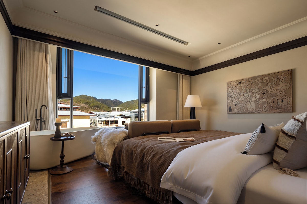 Una habitación de hotel con una cama grande, vistas a edificios y colinas, una lámpara y un cuadro en la pared.