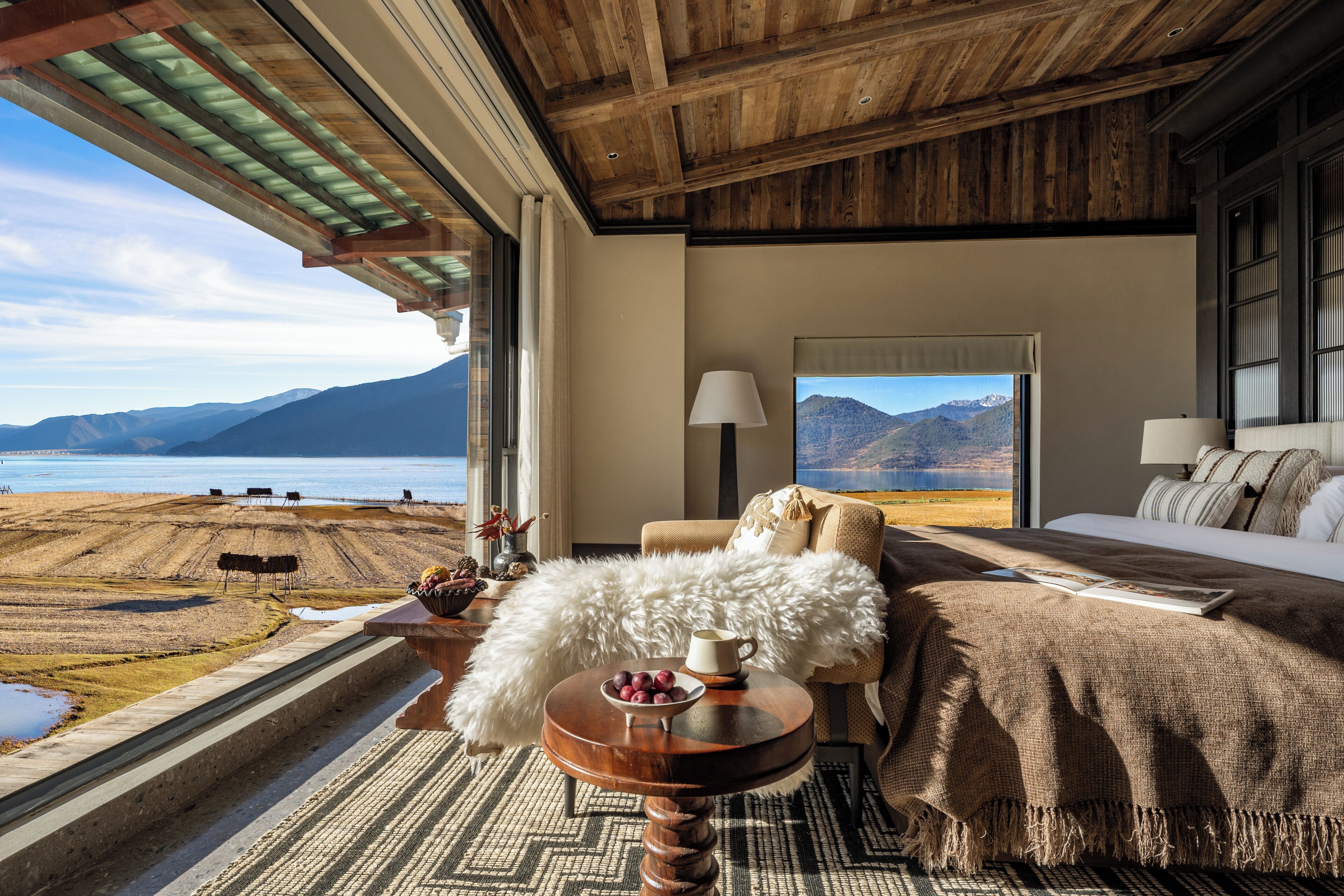 Una habitación con una cama, una mesa con un frutero, una lámpara y vistas a un lago y montañas.