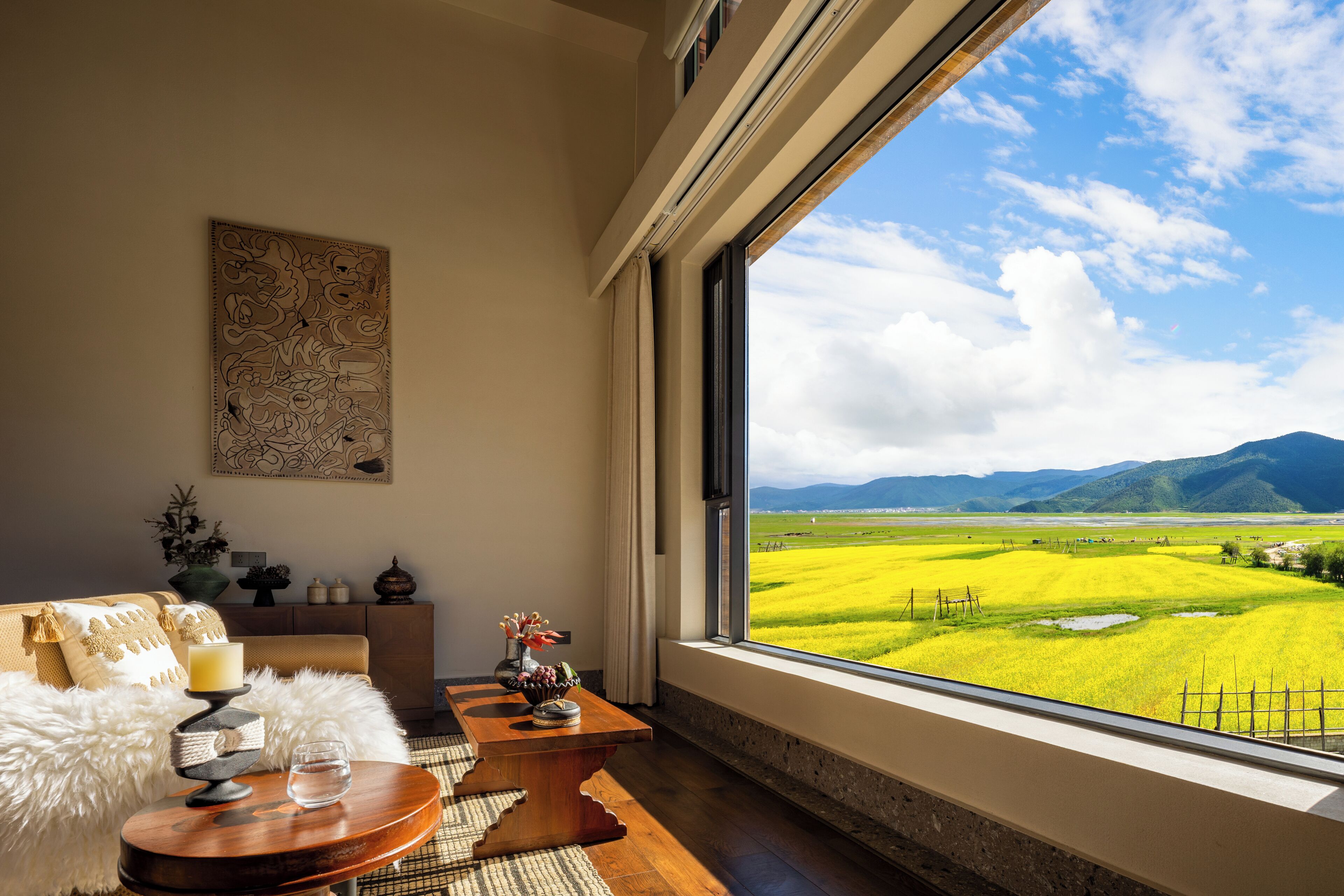 Una habitación con un ventanal que da a un campo, un sofá, una mesa con un jarrón y una pared con un adorno.